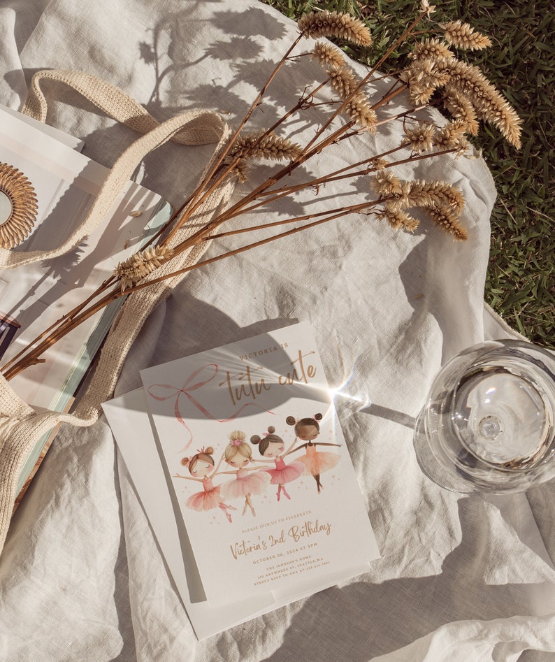 May include: A white invitation card with a pink ribbon and the text "Victoria's 2nd Birthday" in gold lettering. The card features an illustration of four ballerinas in pink tutus and sparkly shoes.