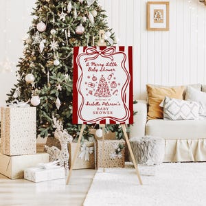 May include: A red and white sign on a wooden easel, with the text "A Merry Little Baby Shower" and a Christmas tree illustration. The sign is in front of a decorated Christmas tree and gifts.