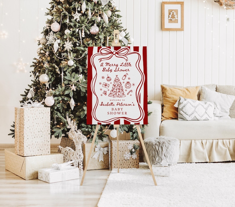 May include: A red and white sign on a wooden easel, with the text "A Merry Little Baby Shower" and a Christmas tree illustration. The sign is in front of a decorated Christmas tree and gifts.