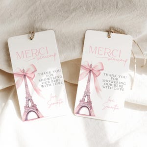 May include: Two white favor tags with pink ribbon and Eiffel Tower design. The text on the tags reads "Merci beaucoup" and "Thank you for showering our bebe with love".