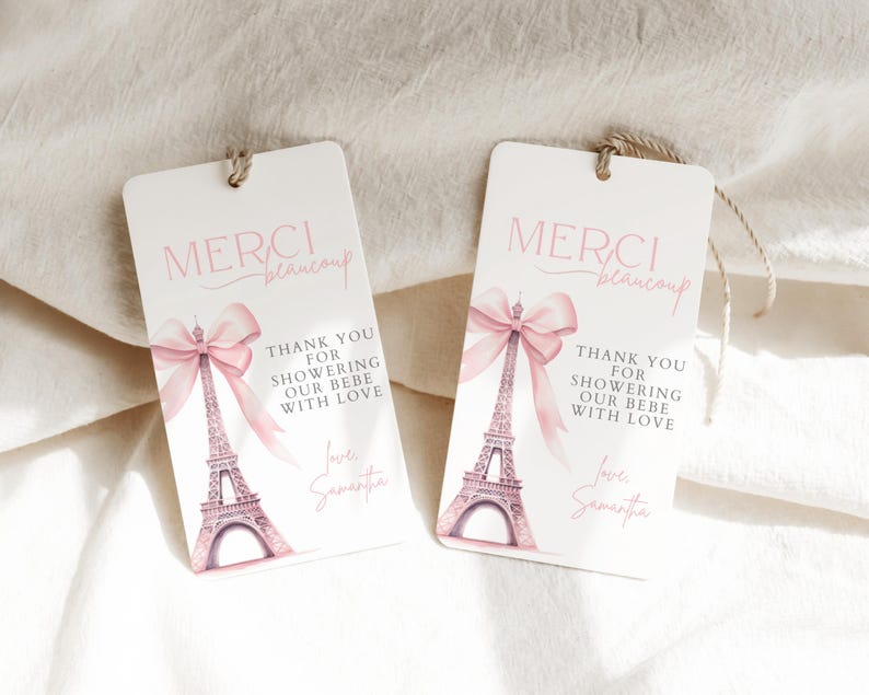 May include: Two white favor tags with pink ribbon and Eiffel Tower design. The text on the tags reads "Merci beaucoup" and "Thank you for showering our bebe with love".