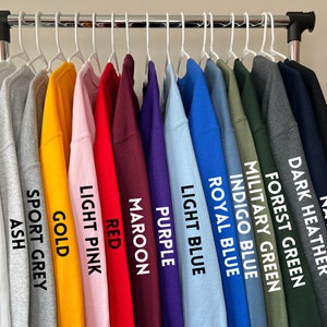 May include: A rack of 15 sweatshirts in various colors, including white, ash, sport grey, gold, light pink, red, maroon, purple, light blue, royal blue, indigo blue, military green, forest green, dark heather, navy, and black.