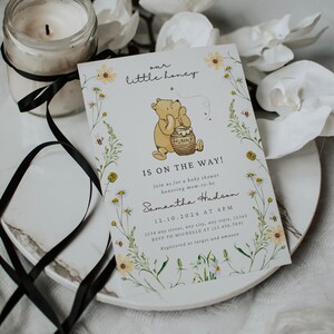 May include: A baby shower invitation with a Winnie the Pooh theme. The invitation features a cartoon bear holding a honey pot, surrounded by floral illustrations. The text reads "our little honey" and "IS ON THE WAY!"