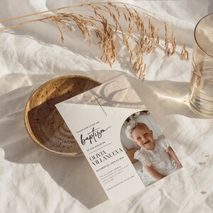 May include: A white baptism invitation with a photo of a baby girl in a white dress. The invitation includes the text "Please join us for the baptism of our daughter Olivia Villanueva, October 06, 2024 at 3PM, The All Saint Church, 123 Anywhere St. Seattle WA, Kindly RSVP to Ana at 1234567890, Lunch Reception to Follow."