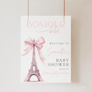 May include: A pink and white baby shower sign with a pink bow on top of the Eiffel Tower. The sign says "Bonjour Bibi" and "Welcome to Samantha's Baby Shower 06.03.24 We're so glad you're here."