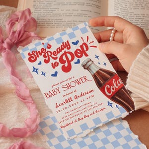 Puede incluir: Invitaci&oacute;n a un baby shower con un dise&ntilde;o retro. La invitaci&oacute;n presenta el texto "She's Ready to Pop" en rojo, junto con una ilustraci&oacute;n de una botella de cola. El fondo tiene un patr&oacute;n de cuadros azules y blancos.