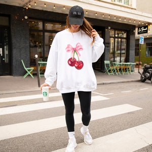 May include: White sweatshirt with a pink bow and two red cherries on the front. The model is wearing a black baseball cap with the text "11:11" on it.