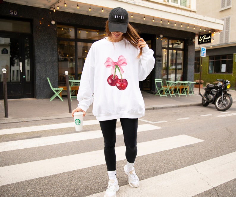 May include: White sweatshirt with a pink bow and two red cherries on the front. The model is wearing a black baseball cap with the text "11:11" on it.