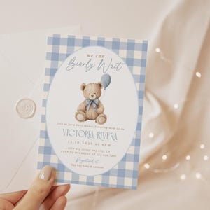 May include: Baby shower invitation with a blue gingham pattern border. The invitation features a watercolor illustration of a teddy bear holding a balloon. The text reads "We can Bearly Wait" and includes event details.
