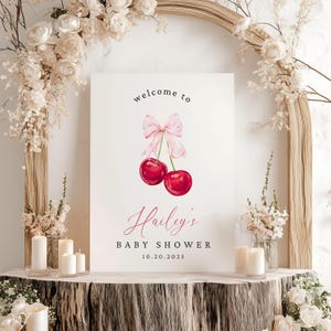 May include: A white welcome sign with a pink bow and two red cherries. The sign reads "Welcome to Hailey's Baby Shower 10.20.2025".