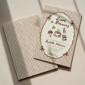 Peut inclure: Deux faire-part de baby shower avec un fond &agrave; rayures. L'invitation pr&eacute;sente le texte "Love is Brewing" et les d&eacute;tails de l'&eacute;v&eacute;nement &agrave; l'encre marron, dont le nom Isabella Peterson et la date.