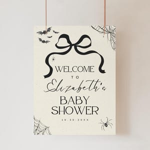 K&ouml;nnte beinhalten: Ein cremefarbenes Schild mit schwarzem Text und Illustrationen, das an einem d&uuml;nnen Faden aufgeh&auml;ngt ist. Auf dem Schild steht "WELCOME to Elizabeth's BABY SHOWER" mit einem Schleifen- und Banddesign. Flederm&auml;use, Spinnen und Spinnweben verzieren das Schild.