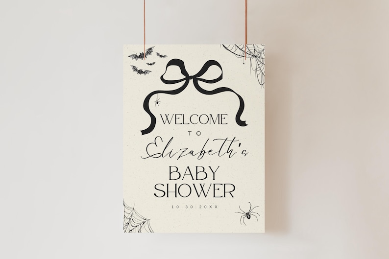K&ouml;nnte beinhalten: Ein cremefarbenes Schild mit schwarzem Text und Illustrationen, das an einem d&uuml;nnen Faden aufgeh&auml;ngt ist. Auf dem Schild steht "WELCOME to Elizabeth's BABY SHOWER" mit einem Schleifen- und Banddesign. Flederm&auml;use, Spinnen und Spinnweben verzieren das Schild.