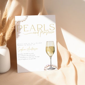 May include: A white invitation card with gold text that reads "Pearls and Prosecco" and a watercolor illustration of a glass of champagne and pearls. The invitation is for a bridal shower in honor of Sophia Anderson. The date and time of the shower are listed as Saturday, October 25, 2023 at four o'clock in the afternoon. The address is listed as 123 Anywhere St., Any City, St 12345. The RSVP information is listed as "roup to Sophia at 123&epsilon;&tau;ά&sigma;&theta;&alpha;&iota;".
