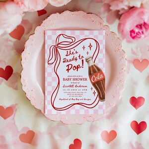 Puede incluir: Una invitaci&oacute;n para un baby shower con un dise&ntilde;o retro. La invitaci&oacute;n presenta el texto "She's Ready to Pop!" y una ilustraci&oacute;n de una botella de cola. La invitaci&oacute;n est&aacute; sobre un fondo a cuadros rosa, rodeada de corazones y flores rosas.