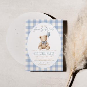 May include: Baby shower invitation with a blue gingham pattern background. The invitation features a watercolor illustration of a teddy bear holding a balloon. The text reads "We can bearly wait" and includes event details.
