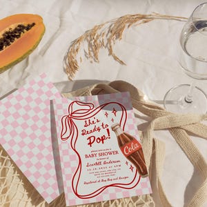 Puede incluir: Invitaci&oacute;n a un baby shower con un patr&oacute;n de cuadros rosas y blancos y el texto "She's Ready to Pop!". Se presenta un gr&aacute;fico de botella de cola vintage. La invitaci&oacute;n est&aacute; sobre una superficie blanca con una papaya y un vaso de agua.