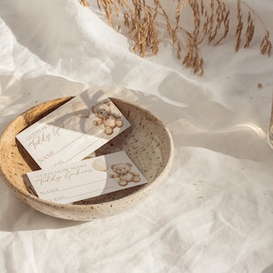 May include: A speckled ceramic bowl holds two baby shower game cards with teddy bear illustrations. The cards read "Guess How Many Teddy Grahams" with spaces for a name and guess. A glass of liquid sits to the right.