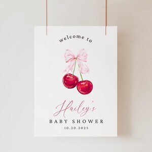 May include: A white sign with the text "welcome to Hailey's Baby Shower 10.20.2025" in black and pink. A pink bow with two red cherries are illustrated above the text.