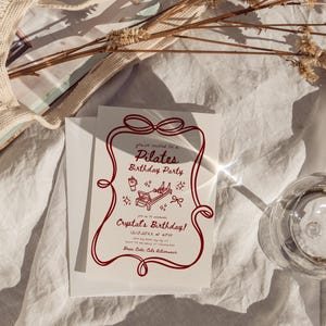 May include: A white invitation with a red border and text that reads "Pilates Birthday Party". The invitation includes illustrations of pilates equipment and a bow. A clear glass and a white envelope are also visible.