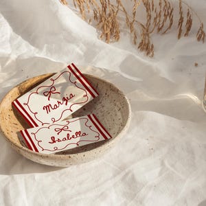 Pu&ograve; includere: Due segnaposto con i nomi "Marzia" e "Isabella" sono in una ciotola di ceramica. I biglietti hanno bordi rossi e disegni a fiocco. La scena &egrave; ambientata su un panno bianco, con un bicchiere d'acqua sullo sfondo.