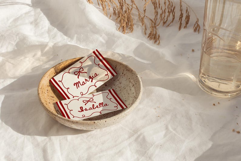 Pu&ograve; includere: Due segnaposto con i nomi "Marzia" e "Isabella" sono in una ciotola di ceramica. I biglietti hanno bordi rossi e disegni a fiocco. La scena &egrave; ambientata su un panno bianco, con un bicchiere d'acqua sullo sfondo.