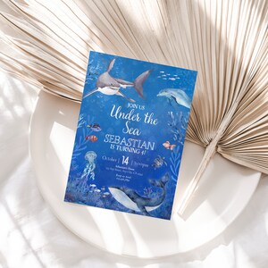 May include: A blue invitation with an underwater theme. The text reads "Join Us Under the Sea, Sebastian is turning 4!" with illustrations of a shark, dolphin, whale, and other sea creatures. The date and time are also included.