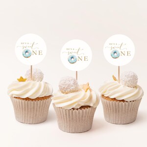 May include: Three white cupcakes with white frosting and gold sprinkles. Each cupcake has a round white topper with blue and gold donut graphic and the text "He's a Sweet One" in gold script.