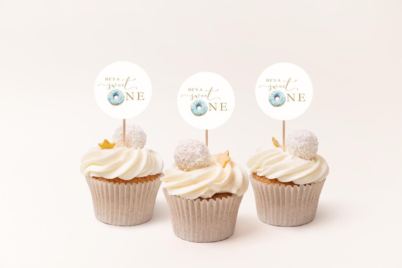 May include: Three white cupcakes with white frosting and gold sprinkles. Each cupcake has a round white topper with blue and gold donut graphic and the text "He's a Sweet One" in gold script.