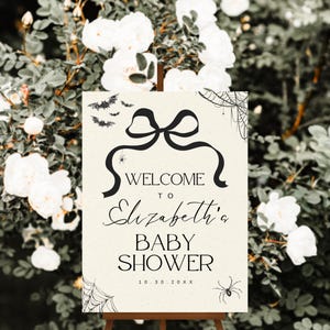 K&ouml;nnte beinhalten: Ein cremefarbenes Willkommensschild f&uuml;r eine Babyparty auf einer Holzstaffelei mit schwarzem Text und dekorativen Elementen. Auf dem Schild steht "Willkommen zu Elizabeth's Baby Shower" mit einem Datum. Flederm&auml;use, Spinnen und Spinnweben zieren die Oberseite und die Ecken.