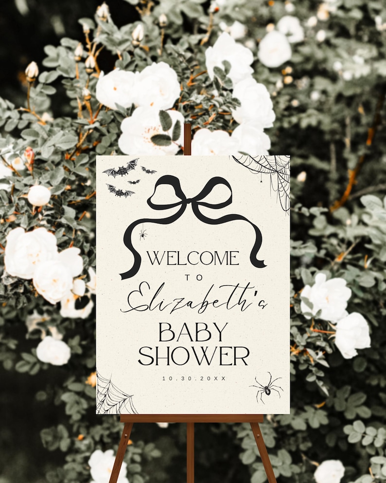 K&ouml;nnte beinhalten: Ein cremefarbenes Willkommensschild f&uuml;r eine Babyparty auf einer Holzstaffelei mit schwarzem Text und dekorativen Elementen. Auf dem Schild steht "Willkommen zu Elizabeth's Baby Shower" mit einem Datum. Flederm&auml;use, Spinnen und Spinnweben zieren die Oberseite und die Ecken.