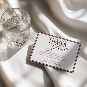 May include: A rectangular white thank you card with dark red border lines and elegant dark red script text that reads Thank You, followed by a message of gratitude and the names Julianna and Benedict, positioned next to a clear glass tumbler.