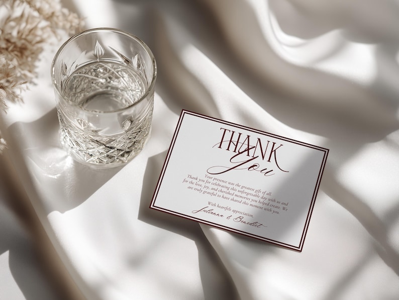 May include: A rectangular white thank you card with dark red border lines and elegant dark red script text that reads Thank You, followed by a message of gratitude and the names Julianna and Benedict, positioned next to a clear glass tumbler.