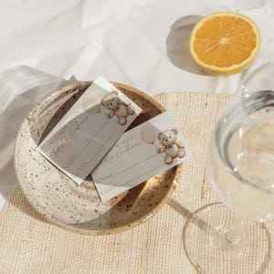 May include: Two baby shower game cards with teddy bear illustrations and the text "Guess How Many Teddy Grahams". The cards are on a speckled ceramic plate, with a half-cut orange and a glass of water.