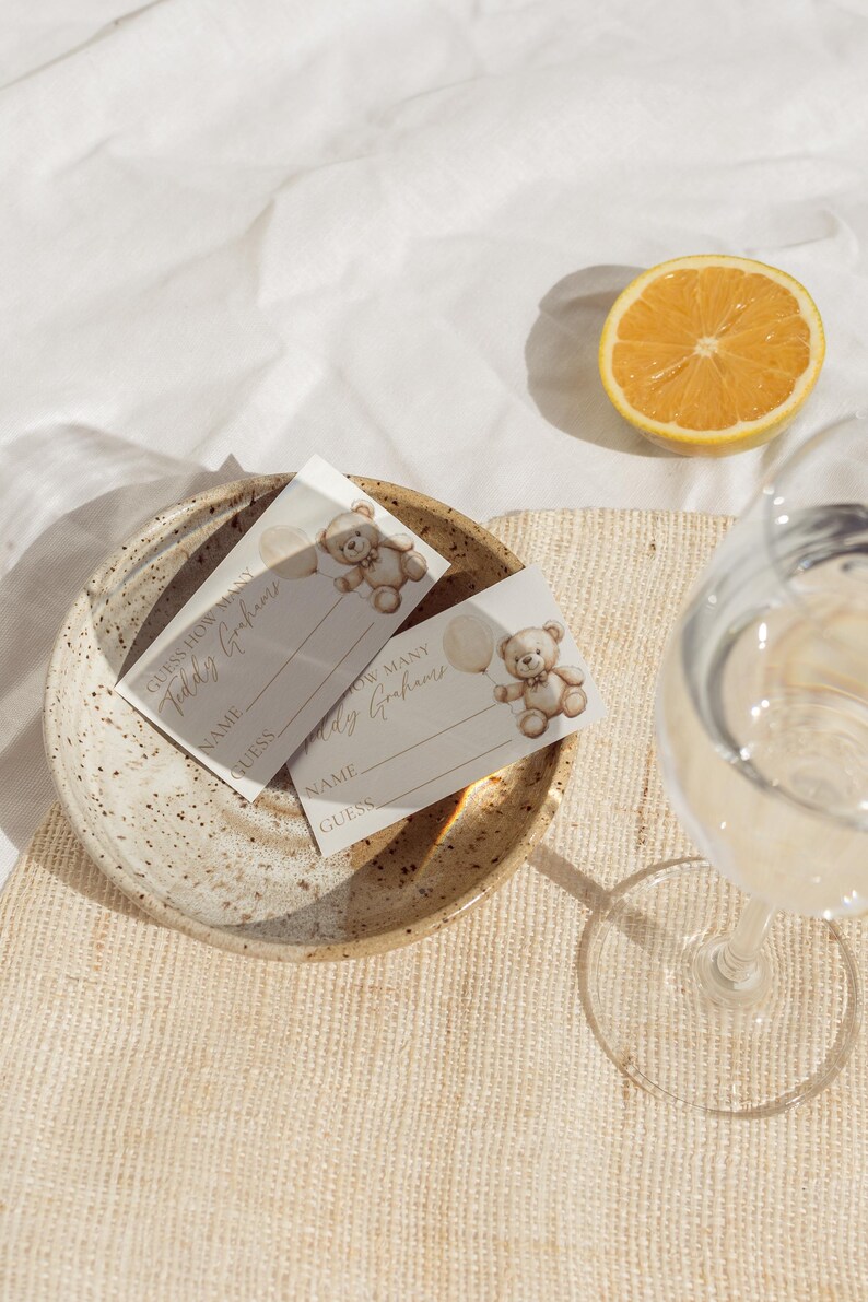 May include: Two baby shower game cards with teddy bear illustrations and the text "Guess How Many Teddy Grahams". The cards are on a speckled ceramic plate, with a half-cut orange and a glass of water.