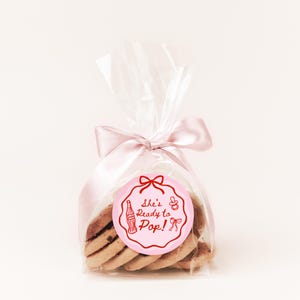 Pode incluir: Uma embalagem transparente de celofane amarrada com uma fita de cetim rosa, contendo v&aacute;rios biscoitos. Uma etiqueta circular rosa com o texto "She's Ready to Pop!" e ilustra&ccedil;&otilde;es com tema de beb&ecirc; est&aacute; anexada &agrave; embalagem.