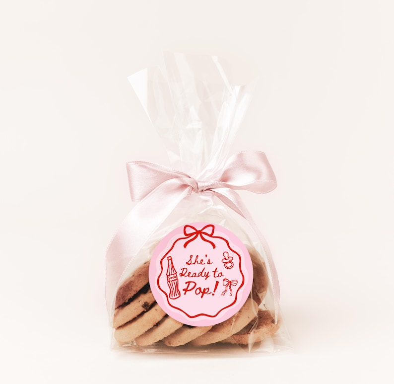 Pode incluir: Uma embalagem transparente de celofane amarrada com uma fita de cetim rosa, contendo v&aacute;rios biscoitos. Uma etiqueta circular rosa com o texto "She's Ready to Pop!" e ilustra&ccedil;&otilde;es com tema de beb&ecirc; est&aacute; anexada &agrave; embalagem.