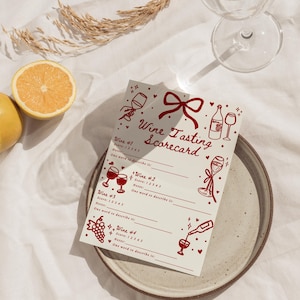 Pu&ograve; includere: Un segnapunti per la degustazione di vini con illustrazioni rosse di bicchieri, bottiglie e un fiocco. Il testo "Wine Tasting Scorecard" &egrave; in alto. La scheda &egrave; su un piatto di ceramica con un limone e un bicchiere d'acqua sullo sfondo.