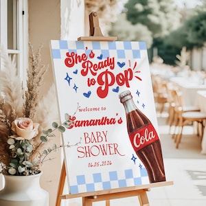 Może przedstawiać: Znak baby shower na sztaludze. Znak ma niebiesko-białe kratkowane tło z czerwonym tekstem, kt&oacute;ry brzmi "She's Ready to Pop" i "Welcome to Samantha's Baby Shower". Prezentowana jest ilustracja butelki coli.