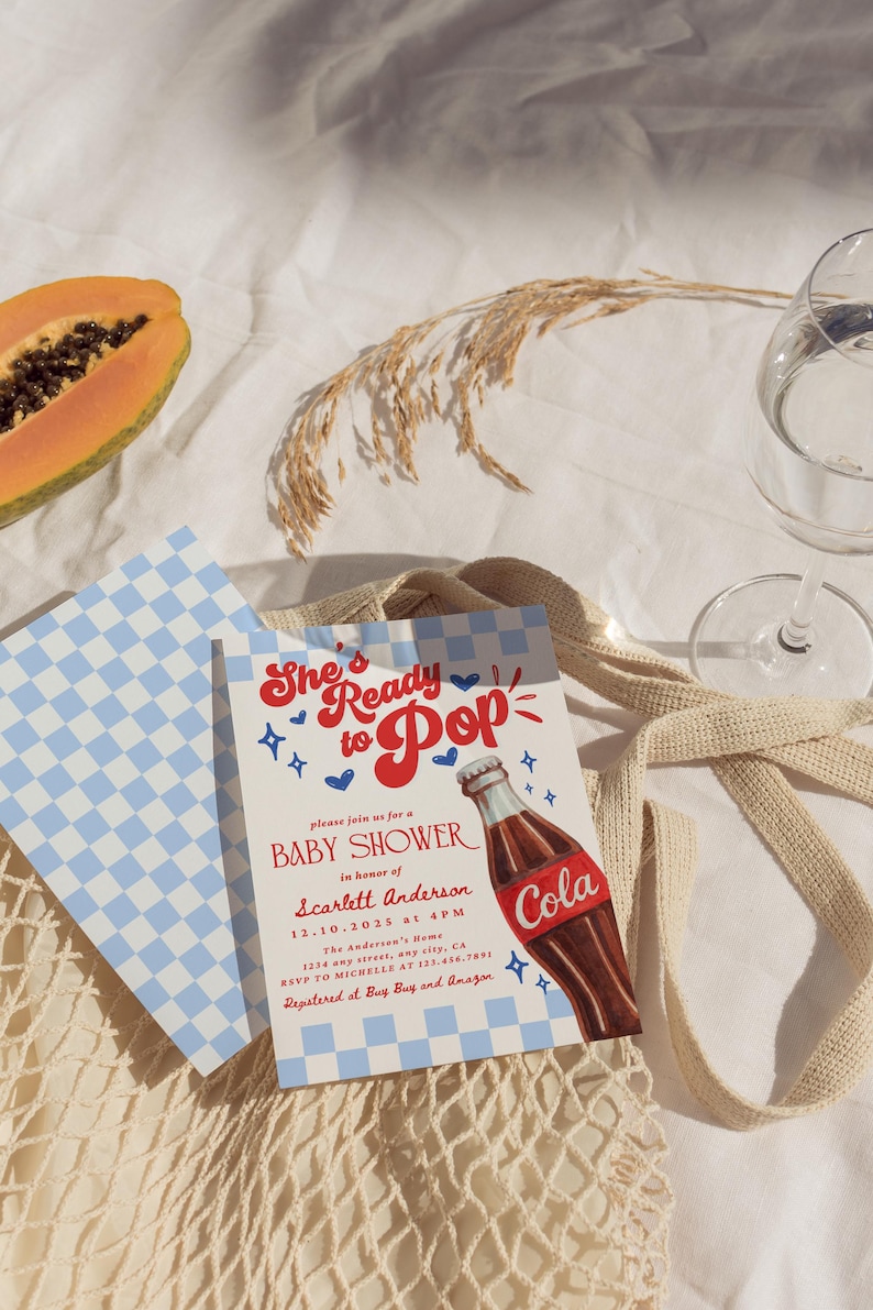 Puede incluir: Invitaci&oacute;n a un baby shower con el texto "She's Ready to Pop" y una ilustraci&oacute;n de una botella de cola. La invitaci&oacute;n est&aacute; sobre un fondo blanco con detalles a cuadros azules. Tambi&eacute;n se ve una rodaja de papaya y un vaso de agua.