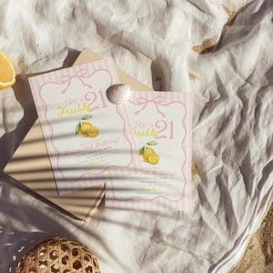 May include: Two rectangular birthday invitation cards featuring a pink striped border, a bow illustration, and yellow lemon graphics. The text reads She's Freshly 21, Birthday, and includes placeholder details. A small white seashell rests on the cards.