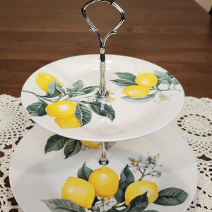 May include: Two-tiered serving stand with a silver handle. The stand is decorated with a lemon tree design featuring yellow lemons, green leaves, and white blossoms.