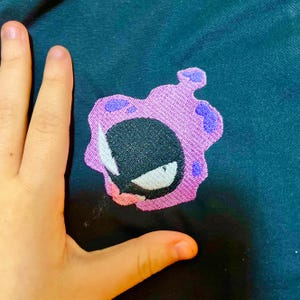 Könnte beinhalten: Gestickter Aufnäher mit einem Cartoon-Geist mit schwarzem Körper, weißem Auge und einem lila wolkenartigen Umriss mit dunkleren lila Akzenten. Der Aufnäher ist auf einen dunkelblauen Stoff genäht.
