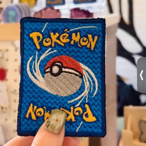 May include: Embroidered patch featuring the back of a Pokémon trading card. The patch has a blue background with yellow text that reads "Pokémon" and a red and white Poké Ball design in the center. The patch is rectangular with a dark blue border.