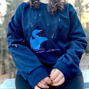 May include: A navy blue sweatshirt with a loon and stars design. The loon is blue and is swimming in a body of water. The stars are yellow.