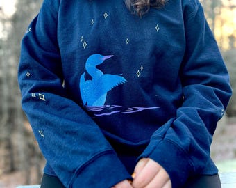May include: A navy blue sweatshirt with a loon and stars design. The loon is blue and is swimming in a body of water. The stars are yellow.