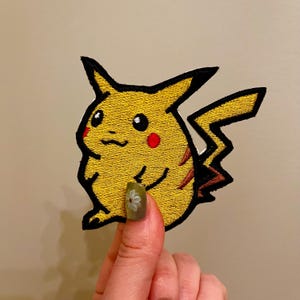 Puede incluir: Parche bordado de Pikachu amarillo con contorno negro, mejillas rojas y cola en zigzag. El parche está sujeto por una mano con un diseño floral en la uña. Ideal para añadir a ropa o accesorios.
