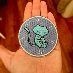 May include: A circular embroidered patch featuring a light blue creature with a long tail and closed eyes centered on a medium blue background. The design includes small white stitched dashes surrounding the figure, all enclosed by a white border.