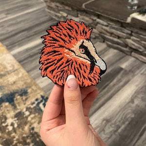 Puede incluir: Parche bordado con una ilustración detallada de la cabeza de un pájaro, que muestra plumas de color naranja y negro vibrantes. El pico del pájaro es blanco y gris, con un ojo negro. El parche es para ropa o accesorios.