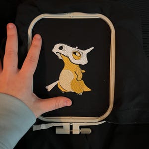 May include: Black fabric with an embroidered Cubone design in yellow, white, and tan. The embroidery is framed by a white embroidery hoop. The design features a cartoon character with a skull mask.